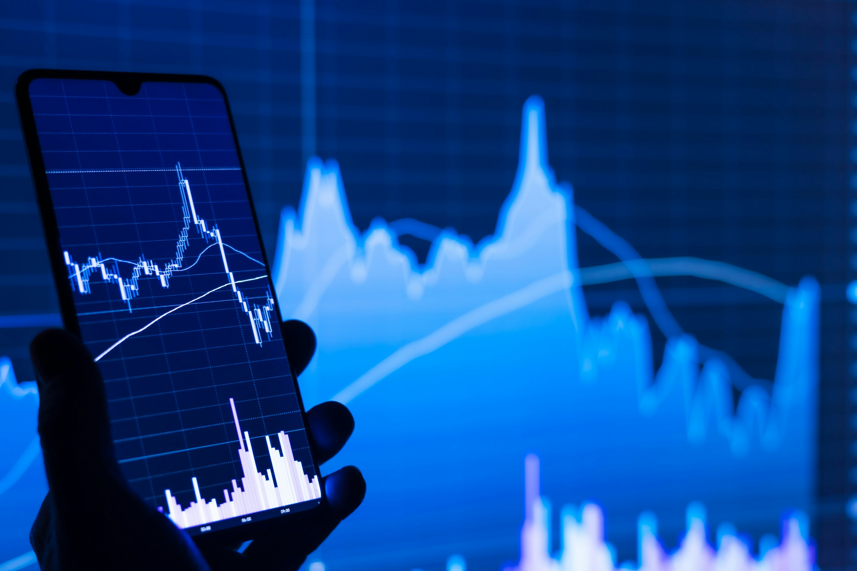 A hand holding a phone displaying financial market charts against a larger data screen